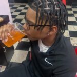 Man with square-sectioned box braids drinking from a plastic cup in a barbershop.
