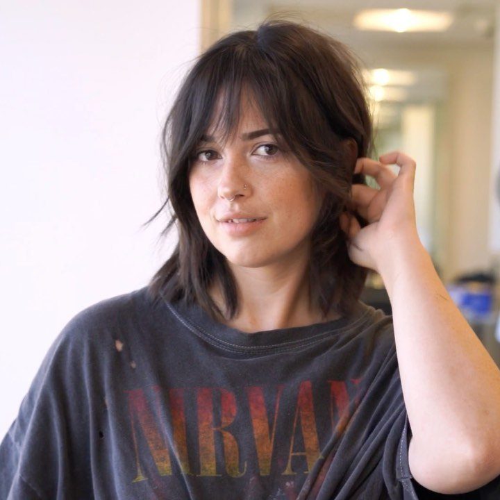 Woman with a medium length brown shag haircut and bangs wearing a grey t-shirt