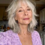 Older woman with layered silver-grey hair and bangs wearing a purple floral blouse.