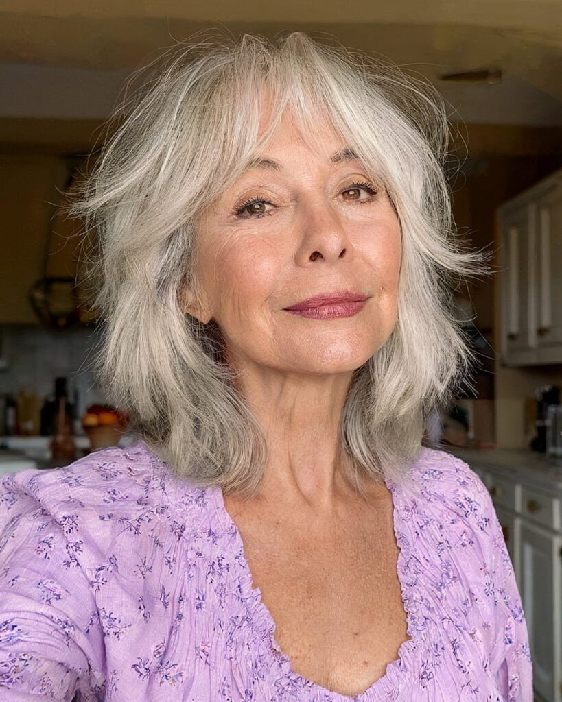 Older woman with layered silver-grey hair and bangs wearing a purple floral blouse.